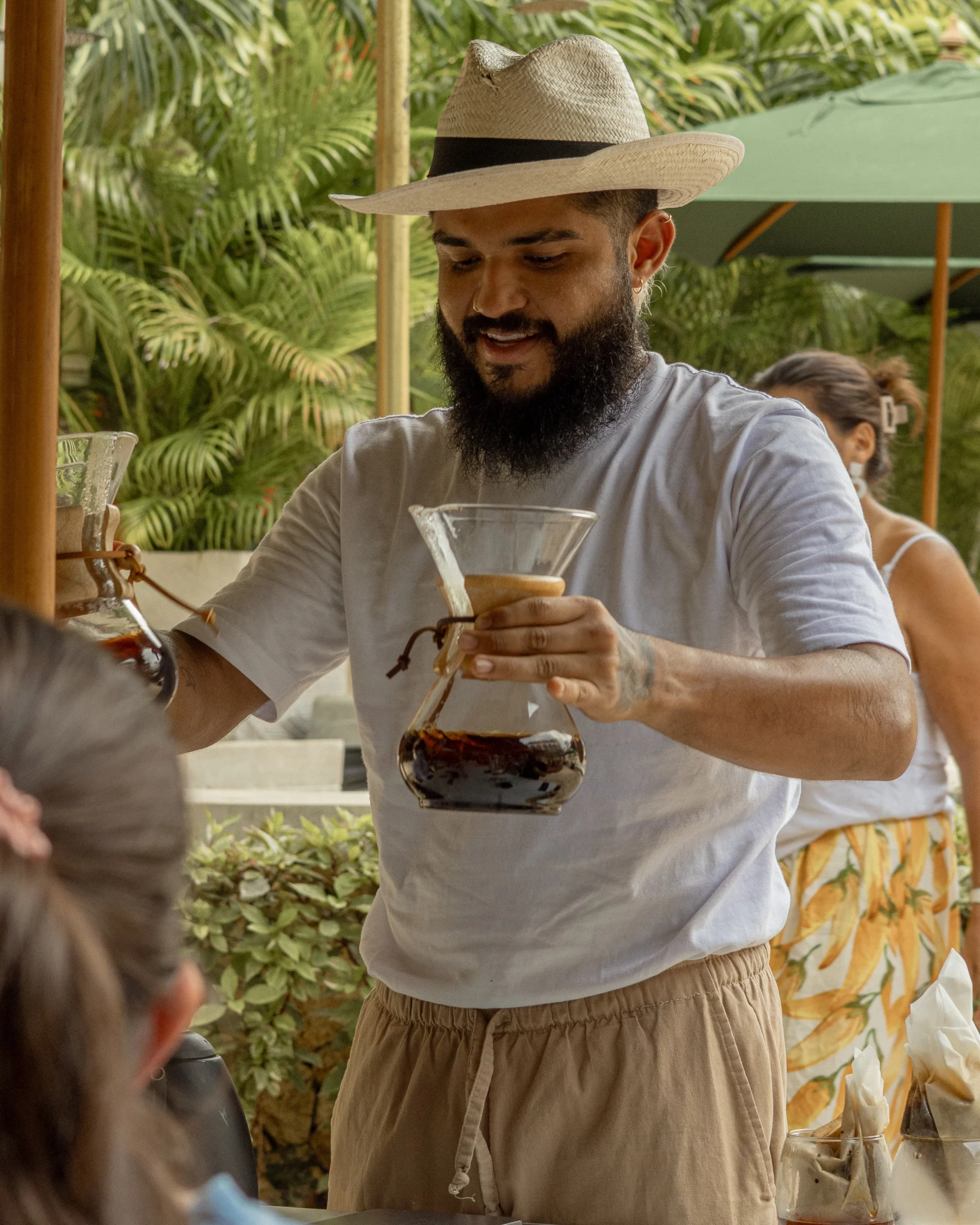 Persona sonriente sosteniendo una cafetera de vidrio con café recién preparado durante una cata al aire libre, con vegetación tropical y otras personas alrededor.