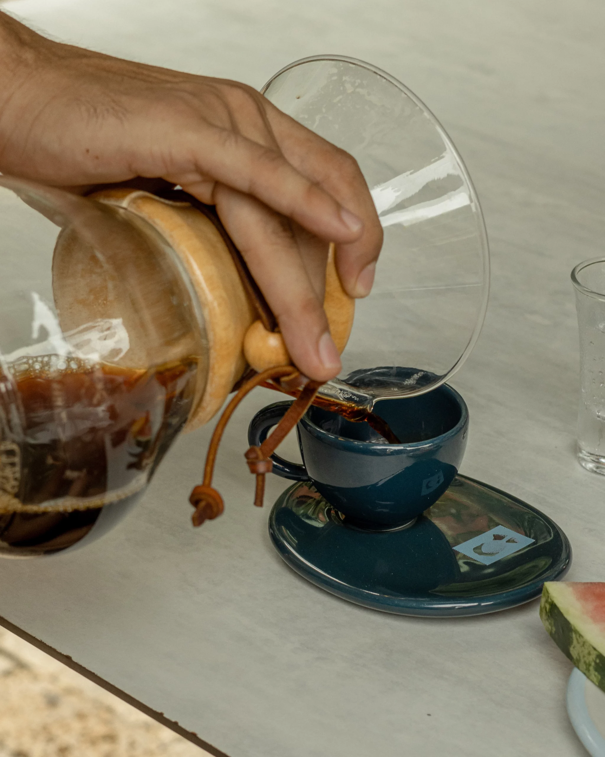 Mano vertiendo café filtrado desde una cafetera de vidrio con cuello de madera hacia una taza azul sobre un platillo, en una mesa clara al aire libre.