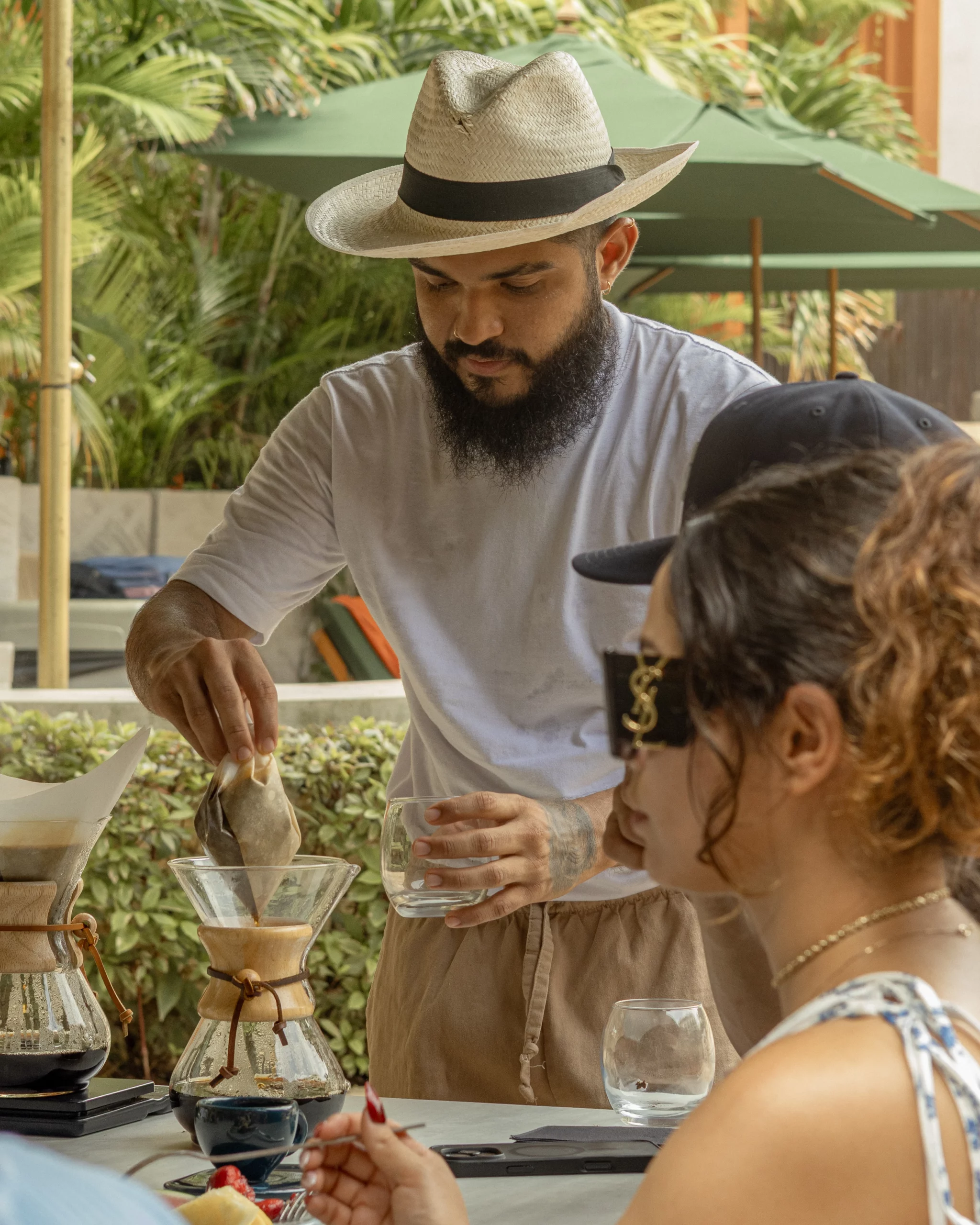 Persona preparando café con método pour-over en una mesa exterior, rodeada de otras personas sentadas; plantas tropicales y sombrillas verdes al fondo.