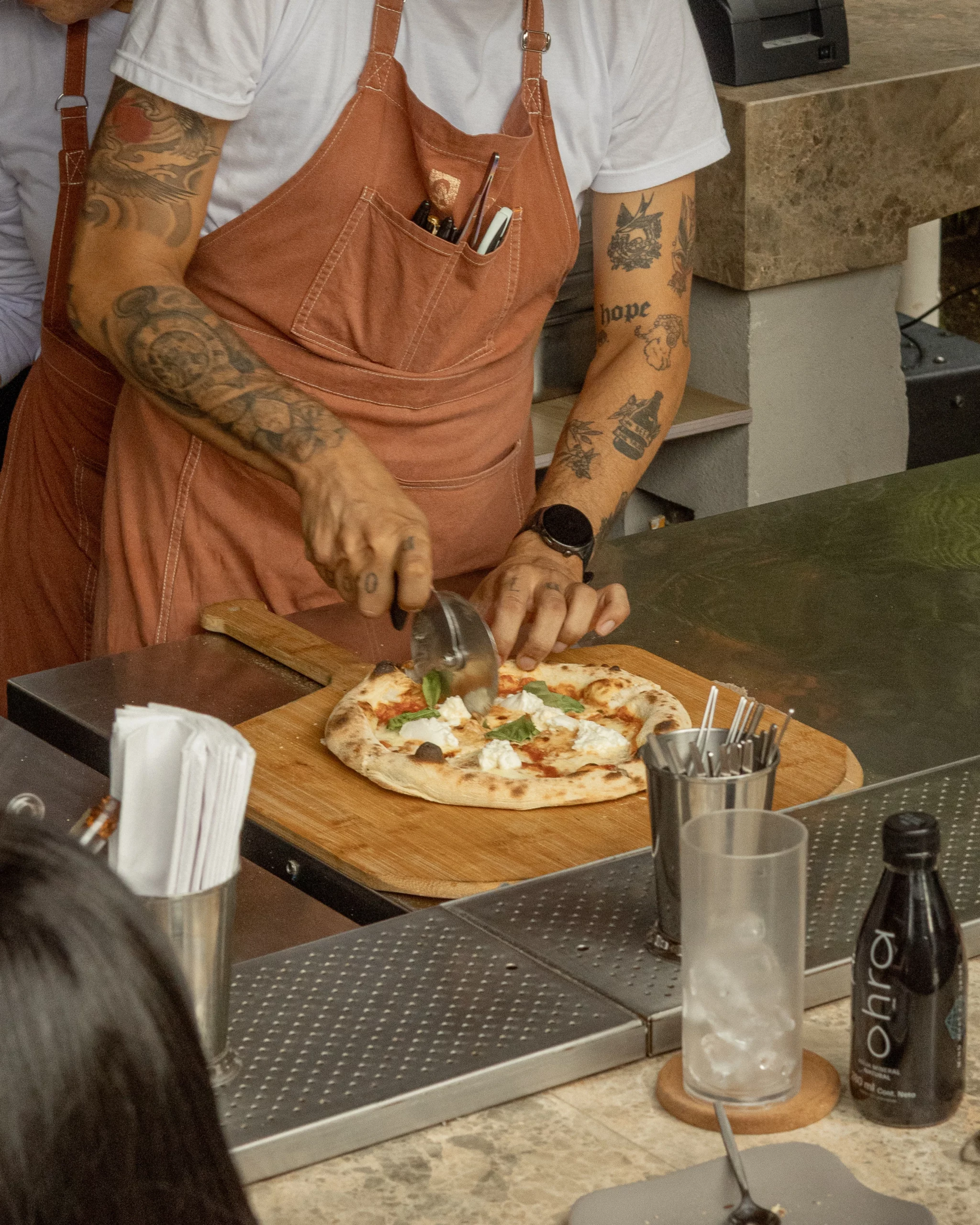 Cook slicing a freshly baked pizza topped with tomato sauce, cheese, and basil on a wooden board at a kitchen counter.