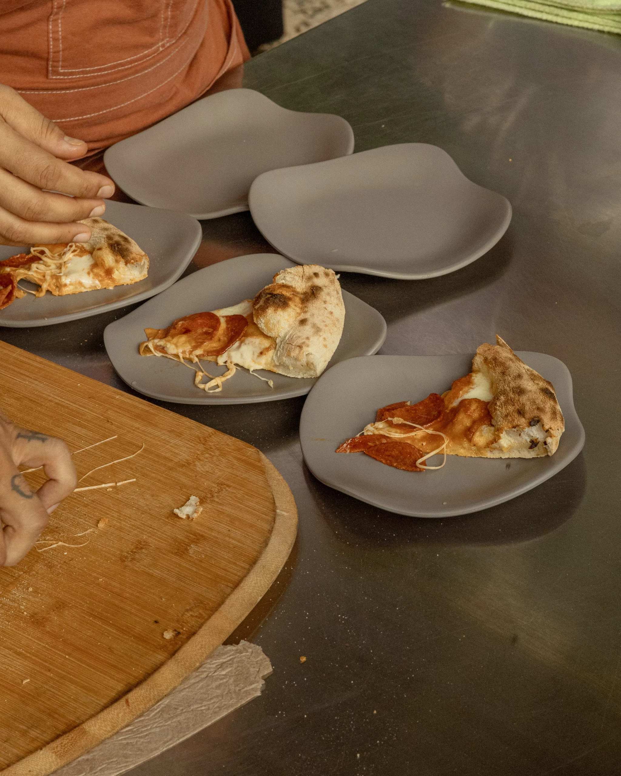 Slices of freshly baked pizza placed on gray plates over a stainless-steel counter, with hands arranging the portions.